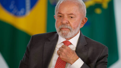 Um homem de cabelos grisalhos e barba, vestindo um terno escuro e uma gravata vermelha com padrões. Ele está sentado, com uma expressão pensativa, enquanto ajusta a gravata. Ao fundo, há bandeiras do Brasil, uma em destaque com as cores verde, amarelo e azul.