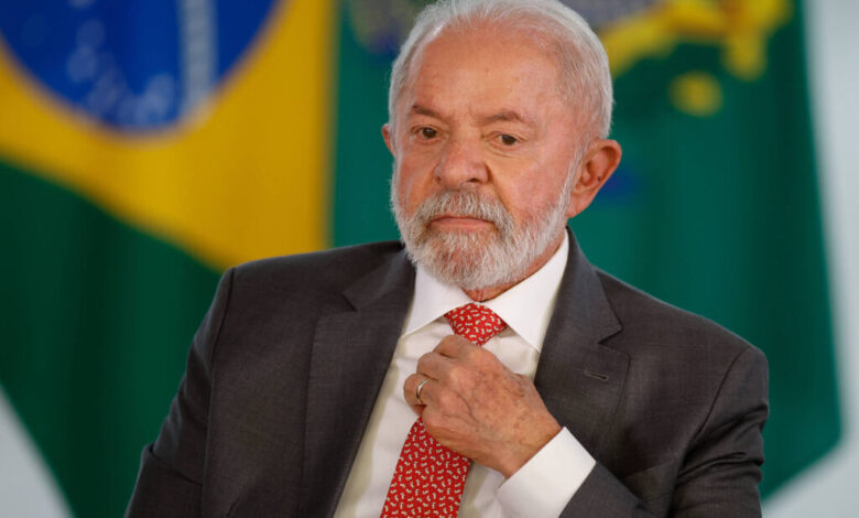 Um homem de cabelos grisalhos e barba, vestindo um terno escuro e uma gravata vermelha com padrões. Ele está sentado, com uma expressão pensativa, enquanto ajusta a gravata. Ao fundo, há bandeiras do Brasil, uma em destaque com as cores verde, amarelo e azul.