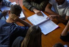 A imagem mostra um grupo de pessoas sentadas ao redor de uma mesa, concentradas em um livro aberto. Algumas mãos estão apontando para o conteúdo do livro, enquanto outras estão apoiadas na mesa. A iluminação é suave, e o ambiente parece ser de estudo ou reunião.