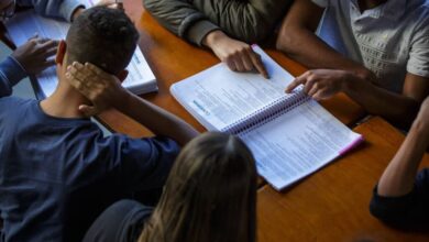 A imagem mostra um grupo de pessoas sentadas ao redor de uma mesa, concentradas em um livro aberto. Algumas mãos estão apontando para o conteúdo do livro, enquanto outras estão apoiadas na mesa. A iluminação é suave, e o ambiente parece ser de estudo ou reunião.