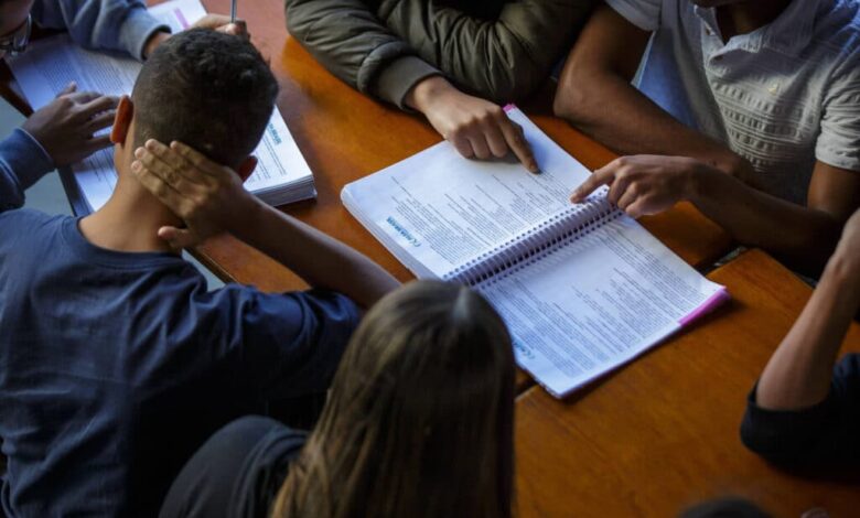 A imagem mostra um grupo de pessoas sentadas ao redor de uma mesa, concentradas em um livro aberto. Algumas mãos estão apontando para o conteúdo do livro, enquanto outras estão apoiadas na mesa. A iluminação é suave, e o ambiente parece ser de estudo ou reunião.