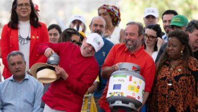 A imagem mostra um evento ao ar livre com várias pessoas reunidas. No centro, um homem de camisa vermelha e chapéu branco segura um botijão de gás, enquanto outro homem ao seu lado, vestido de azul, segura um recipiente semelhante. Ao fundo, há uma multidão de pessoas, algumas usando bonés e chapéus coloridos. O ambiente parece festivo, com sorrisos e interações entre os participantes.