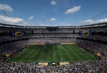 A imagem mostra um estádio de futebol lotado, com torcedores nas arquibancadas. O campo está visível, com jogadores em ação. O céu está parcialmente nublado, e há grandes telões visíveis nas laterais do estádio. A atmosfera é de grande expectativa e emoção, típica de um evento esportivo.