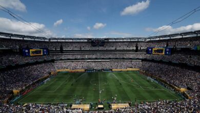 A imagem mostra um estádio de futebol lotado, com torcedores nas arquibancadas. O campo está visível, com jogadores em ação. O céu está parcialmente nublado, e há grandes telões visíveis nas laterais do estádio. A atmosfera é de grande expectativa e emoção, típica de um evento esportivo.