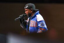 Um artista está se apresentando em um palco, segurando um microfone. Ele usa um boné preto e uma jaqueta colorida com detalhes em azul e vermelho. O fundo é desfocado, destacando o artista em primeiro plano.