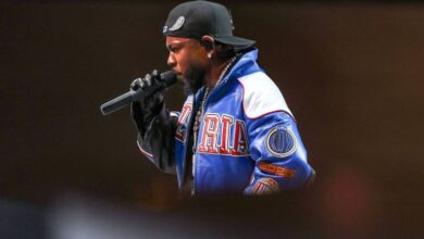 Um artista está se apresentando em um palco, segurando um microfone. Ele usa um boné preto e uma jaqueta colorida com detalhes em azul e vermelho. O fundo é desfocado, destacando o artista em primeiro plano.