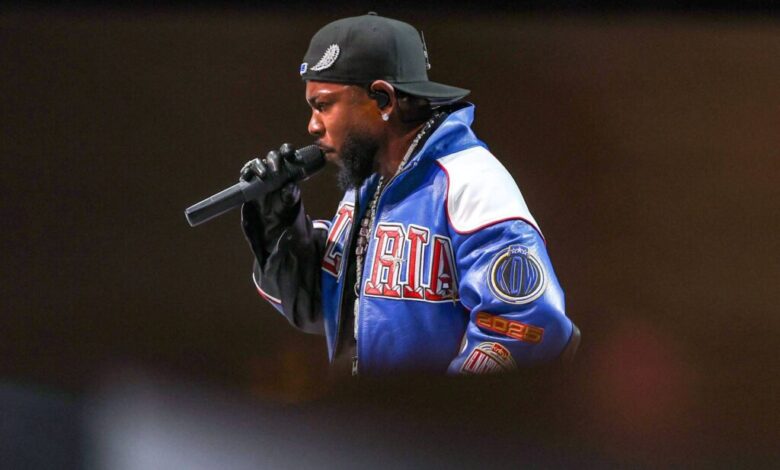 Um artista está se apresentando em um palco, segurando um microfone. Ele usa um boné preto e uma jaqueta colorida com detalhes em azul e vermelho. O fundo é desfocado, destacando o artista em primeiro plano.