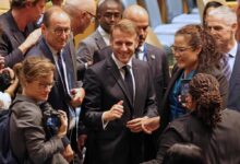 A imagem mostra um grupo de pessoas em um ambiente formal, possivelmente no Conselho de Segurança da ONU. No centro, um homem sorridente, vestido com um terno escuro, está cercado por várias pessoas que parecem estar interagindo com ele. Algumas pessoas estão segurando câmeras, enquanto outras observam. O ambiente é bem iluminado e há cadeiras ao fundo.