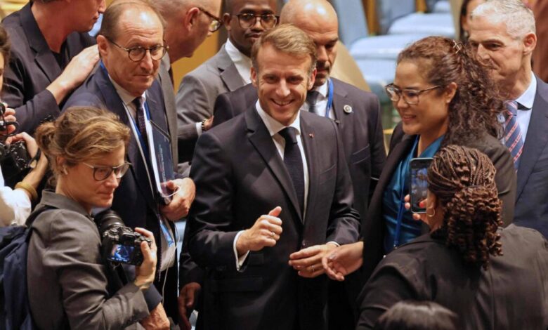 A imagem mostra um grupo de pessoas em um ambiente formal, possivelmente no Conselho de Segurança da ONU. No centro, um homem sorridente, vestido com um terno escuro, está cercado por várias pessoas que parecem estar interagindo com ele. Algumas pessoas estão segurando câmeras, enquanto outras observam. O ambiente é bem iluminado e há cadeiras ao fundo.