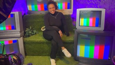 Pessoa sentada em sofá verde cercada por cinco televisores antigos exibindo tela de teste colorida. Ambiente iluminado com luz roxa e decoração com folhas na parede.