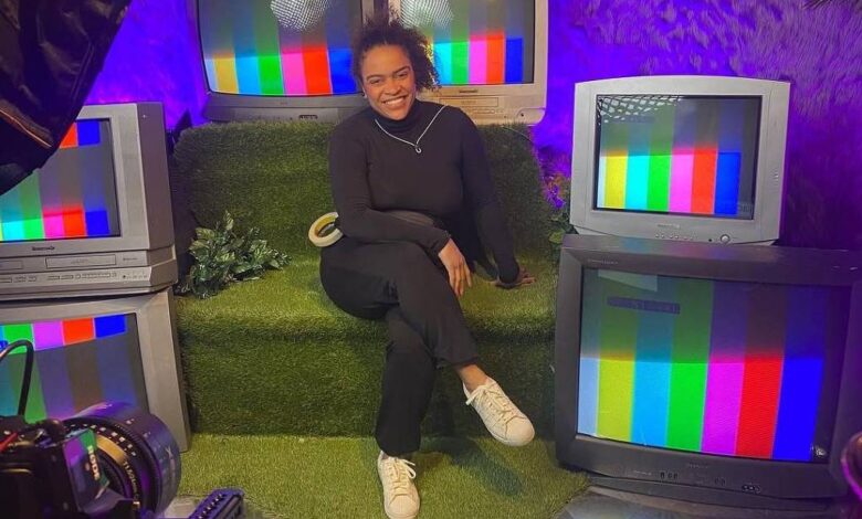 Pessoa sentada em sofá verde cercada por cinco televisores antigos exibindo tela de teste colorida. Ambiente iluminado com luz roxa e decoração com folhas na parede.
