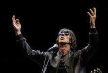 Um artista está se apresentando em um palco, usando óculos escuros e uma jaqueta preta. Ele está com os braços levantados, em uma pose expressiva, enquanto canta em um microfone. Ao fundo, há um cenário escuro, destacando a figura do artista e um violão visível ao lado.
