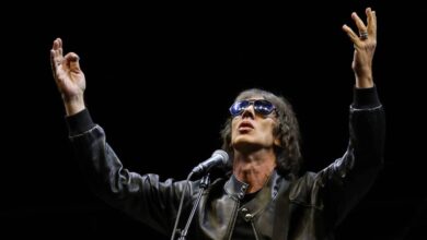 Um artista está se apresentando em um palco, usando óculos escuros e uma jaqueta preta. Ele está com os braços levantados, em uma pose expressiva, enquanto canta em um microfone. Ao fundo, há um cenário escuro, destacando a figura do artista e um violão visível ao lado.