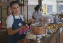 Mulher com avental azul e camiseta branca limpa a vitrine de bolos em balcão de confeitaria. Ao fundo, outra funcionária e mesas externas visíveis.