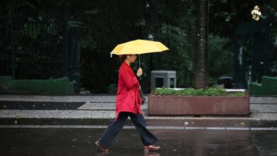 Pessoa vestindo casaco vermelho caminha na calçada molhada segurando um guarda-chuva amarelo aberto. Ambiente urbano com árvores e bancos ao fundo em dia escuro e chuvoso.