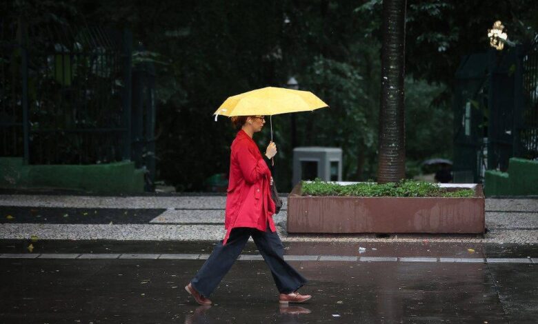 Pessoa vestindo casaco vermelho caminha na calçada molhada segurando um guarda-chuva amarelo aberto. Ambiente urbano com árvores e bancos ao fundo em dia escuro e chuvoso.