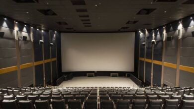 Sala de cinema com fileiras de poltronas pretas vazias voltadas para uma grande tela branca. As paredes laterais são cinza com faixas amarelas e iluminação embutida no teto e nas paredes.