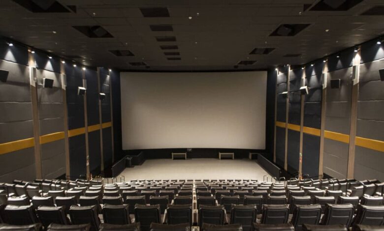 Sala de cinema com fileiras de poltronas pretas vazias voltadas para uma grande tela branca. As paredes laterais são cinza com faixas amarelas e iluminação embutida no teto e nas paredes.