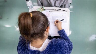 A imagem mostra a parte de trás de uma estudante com cabelo preso em um coque, usando um suéter azul. Ela está escrevendo em uma folha de papel sobre uma mesa, que também contém uma garrafa de água, canetas e outros papéis. O ambiente parece ser uma sala de aula, com piso verde.