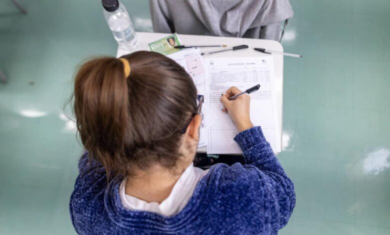 A imagem mostra a parte de trás de uma estudante com cabelo preso em um coque, usando um suéter azul. Ela está escrevendo em uma folha de papel sobre uma mesa, que também contém uma garrafa de água, canetas e outros papéis. O ambiente parece ser uma sala de aula, com piso verde.