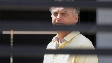 Homem de meia-idade com camisa polo amarela observa à frente, enquadrado por persianas horizontais pretas que criam faixas escuras na imagem.