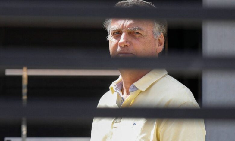 Homem de meia-idade com camisa polo amarela observa à frente, enquadrado por persianas horizontais pretas que criam faixas escuras na imagem.