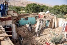 Casa parcialmente destruída com paredes derrubadas e escombros espalhados. Pessoas observam e caminham entre os destroços em área rural com árvores e montanhas ao fundo.