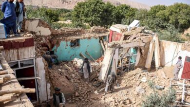 Casa parcialmente destruída com paredes derrubadas e escombros espalhados. Pessoas observam e caminham entre os destroços em área rural com árvores e montanhas ao fundo.