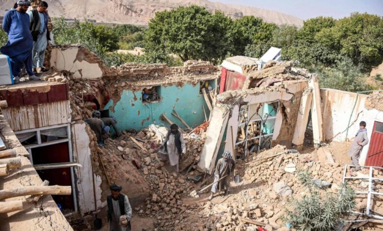 Casa parcialmente destruída com paredes derrubadas e escombros espalhados. Pessoas observam e caminham entre os destroços em área rural com árvores e montanhas ao fundo.