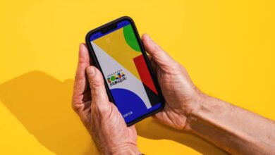 Mãos seguram um celular com tela exibindo formas geométricas coloridas em fundo amarelo. A sombra das mãos e do aparelho aparece à esquerda.