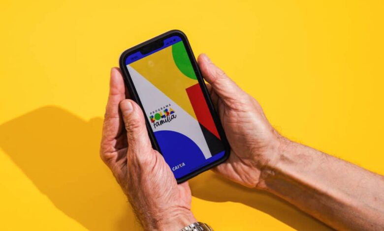 Mãos seguram um celular com tela exibindo formas geométricas coloridas em fundo amarelo. A sombra das mãos e do aparelho aparece à esquerda.