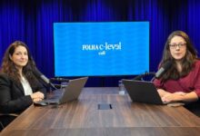 Duas mulheres sentadas em frente a uma mesa de madeira em estúdio com fundo azul escuro. Ambas usam laptops e falam em microfones de mesa. Ao fundo, tela exibe logo