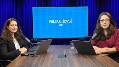 Duas mulheres sentadas em frente a uma mesa de madeira em estúdio com fundo azul escuro. Ambas usam laptops e falam em microfones de mesa. Ao fundo, tela exibe logo