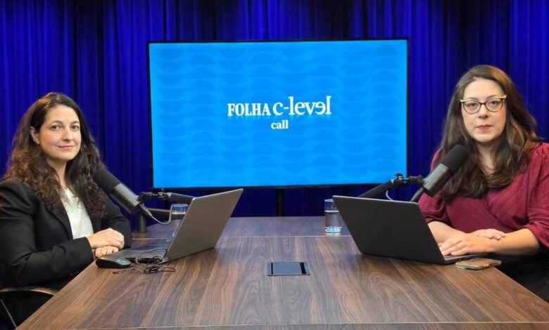 Duas mulheres sentadas em frente a uma mesa de madeira em estúdio com fundo azul escuro. Ambas usam laptops e falam em microfones de mesa. Ao fundo, tela exibe logo