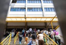 Grupo de pessoas de costas sobe escada com corrimão amarelo em direção à entrada de prédio branco de vários andares com janelas azuis. Estrutura metálica amarela cobre parte da escada.
