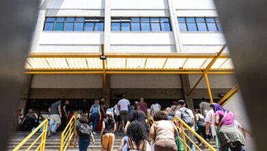 Grupo de pessoas de costas sobe escada com corrimão amarelo em direção à entrada de prédio branco de vários andares com janelas azuis. Estrutura metálica amarela cobre parte da escada.