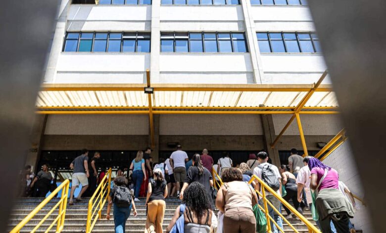 Grupo de pessoas de costas sobe escada com corrimão amarelo em direção à entrada de prédio branco de vários andares com janelas azuis. Estrutura metálica amarela cobre parte da escada.