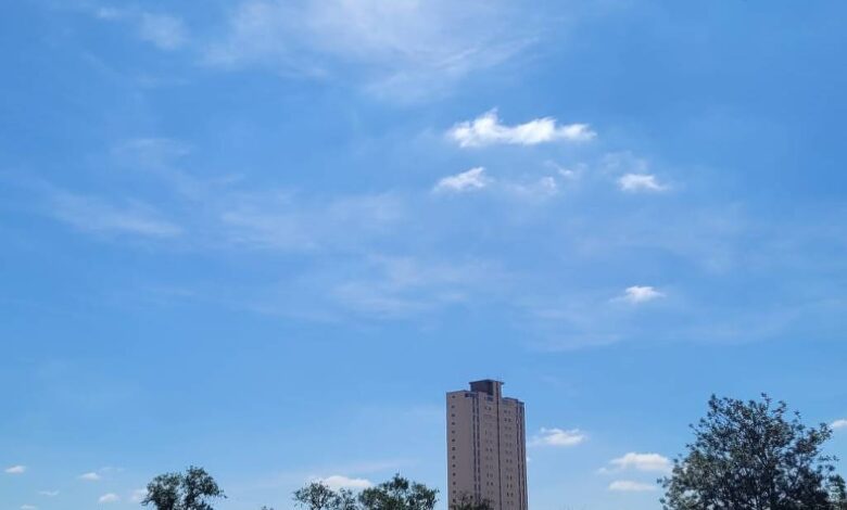 Céu azul claro com poucas nuvens brancas dispersas acima de telhados residenciais e árvores. Um prédio alto aparece ao fundo, sob luz do dia clara.
