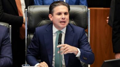 Homem de terno azul escuro e gravata verde senta em cadeira de couro preta, falando ao microfone em ambiente formal, com outras pessoas ao fundo.