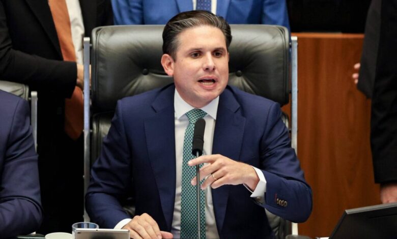 Homem de terno azul escuro e gravata verde senta em cadeira de couro preta, falando ao microfone em ambiente formal, com outras pessoas ao fundo.