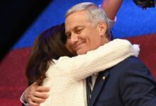 Homem de cabelos grisalhos veste terno azul escuro e sorri com os olhos fechados enquanto é abraçado por mulher de cabelos castanhos e casaco branco. Fundo desfocado com luzes coloridas.