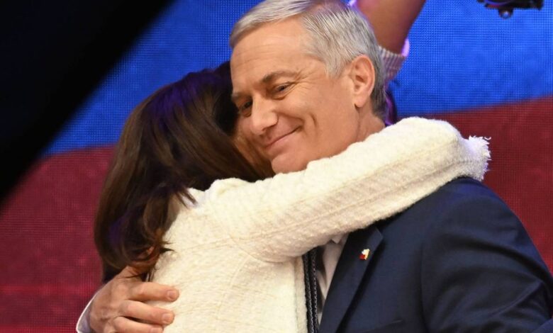 Homem de cabelos grisalhos veste terno azul escuro e sorri com os olhos fechados enquanto é abraçado por mulher de cabelos castanhos e casaco branco. Fundo desfocado com luzes coloridas.