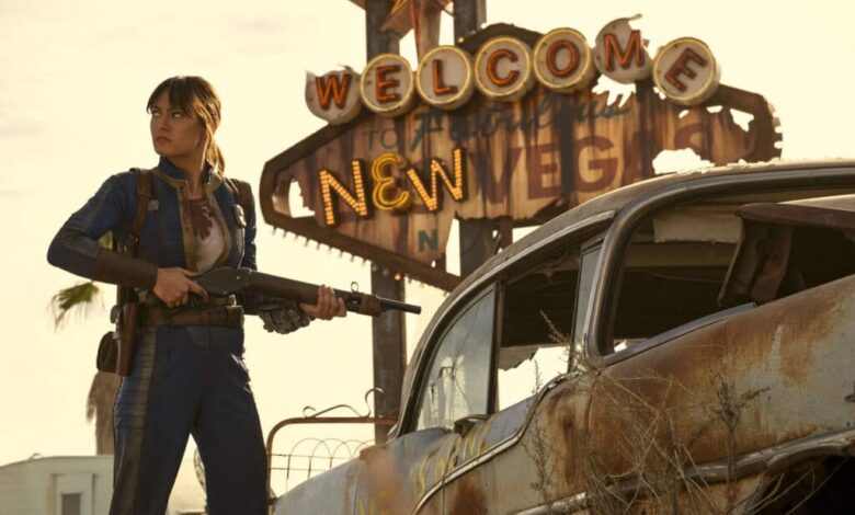 Mulher vestindo macacão azul e segurando rifle está ao lado de carro enferrujado. Ao fundo, letreiro antigo iluminado com as palavras