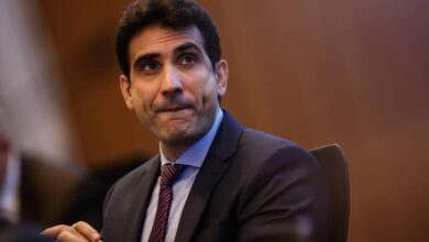 Um homem com cabelo escuro e liso, usando um terno escuro e uma gravata listrada, está sentado em uma mesa. Ele parece pensativo, com a boca levemente entreaberta e as mãos unidas na frente. O fundo é desfocado, sugerindo um ambiente de reunião ou conferência.