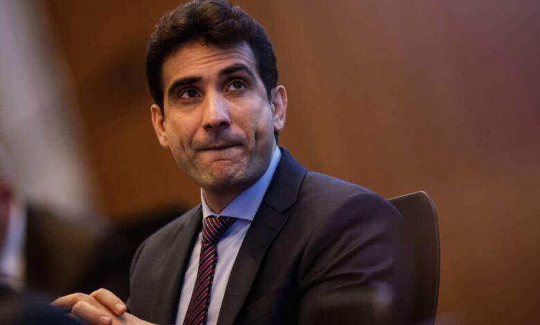 Um homem com cabelo escuro e liso, usando um terno escuro e uma gravata listrada, está sentado em uma mesa. Ele parece pensativo, com a boca levemente entreaberta e as mãos unidas na frente. O fundo é desfocado, sugerindo um ambiente de reunião ou conferência.