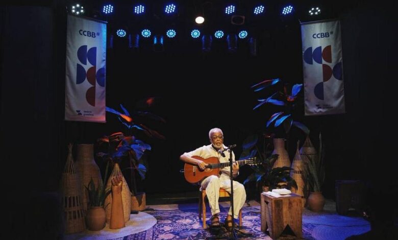 Músico sentado no centro do palco toca violão sob luzes azuis. Dois banners do CCBB pendem ao fundo, com plantas e objetos decorativos ao redor.