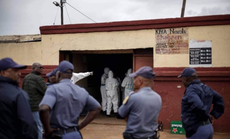 Quatro policiais de uniforme azul estão em primeiro plano, de costas e de perfil, observando a entrada de um prédio com paredes amarelas e vermelhas. Na entrada, três pessoas vestindo roupas brancas de proteção estão dentro do estabelecimento, uma delas apontando para o interior. Acima da porta, há uma placa com texto parcialmente legível indicando