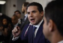 Homem de terno azul e gravata azul fala gesticulando com a mão direita em ambiente interno com outras pessoas ao fundo.