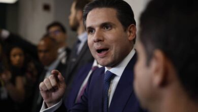 Homem de terno azul e gravata azul fala gesticulando com a mão direita em ambiente interno com outras pessoas ao fundo.