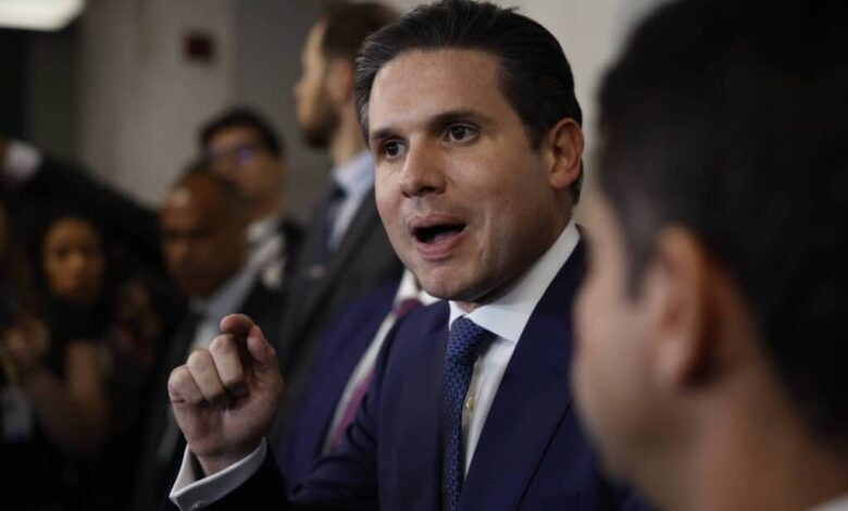 Homem de terno azul e gravata azul fala gesticulando com a mão direita em ambiente interno com outras pessoas ao fundo.